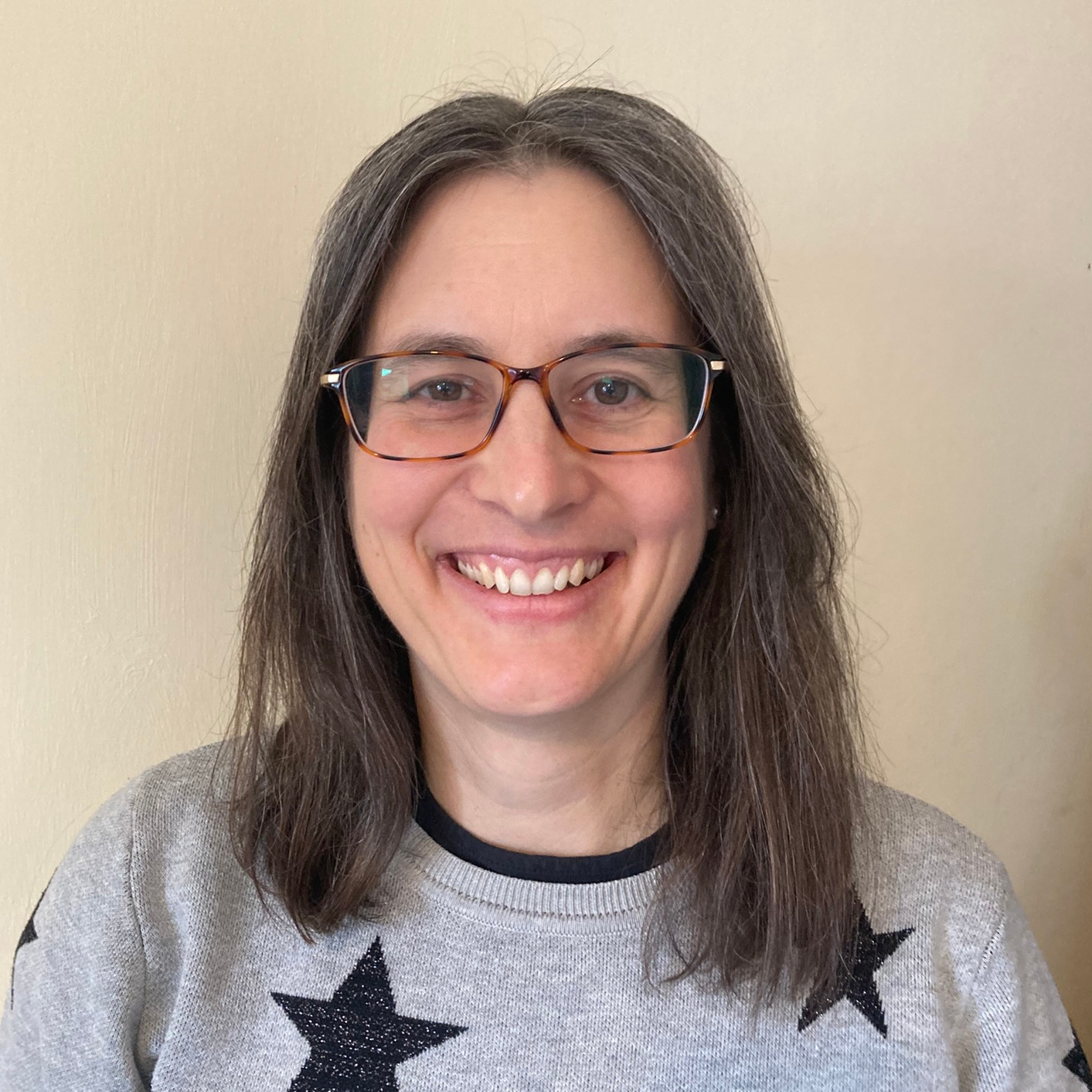 Headshot of Heather, a white middle-aged woman with shoulder-length, dark straight hair and tortoisehell glasses. She has a big smile and is wearing a grey jumper with black stars on.